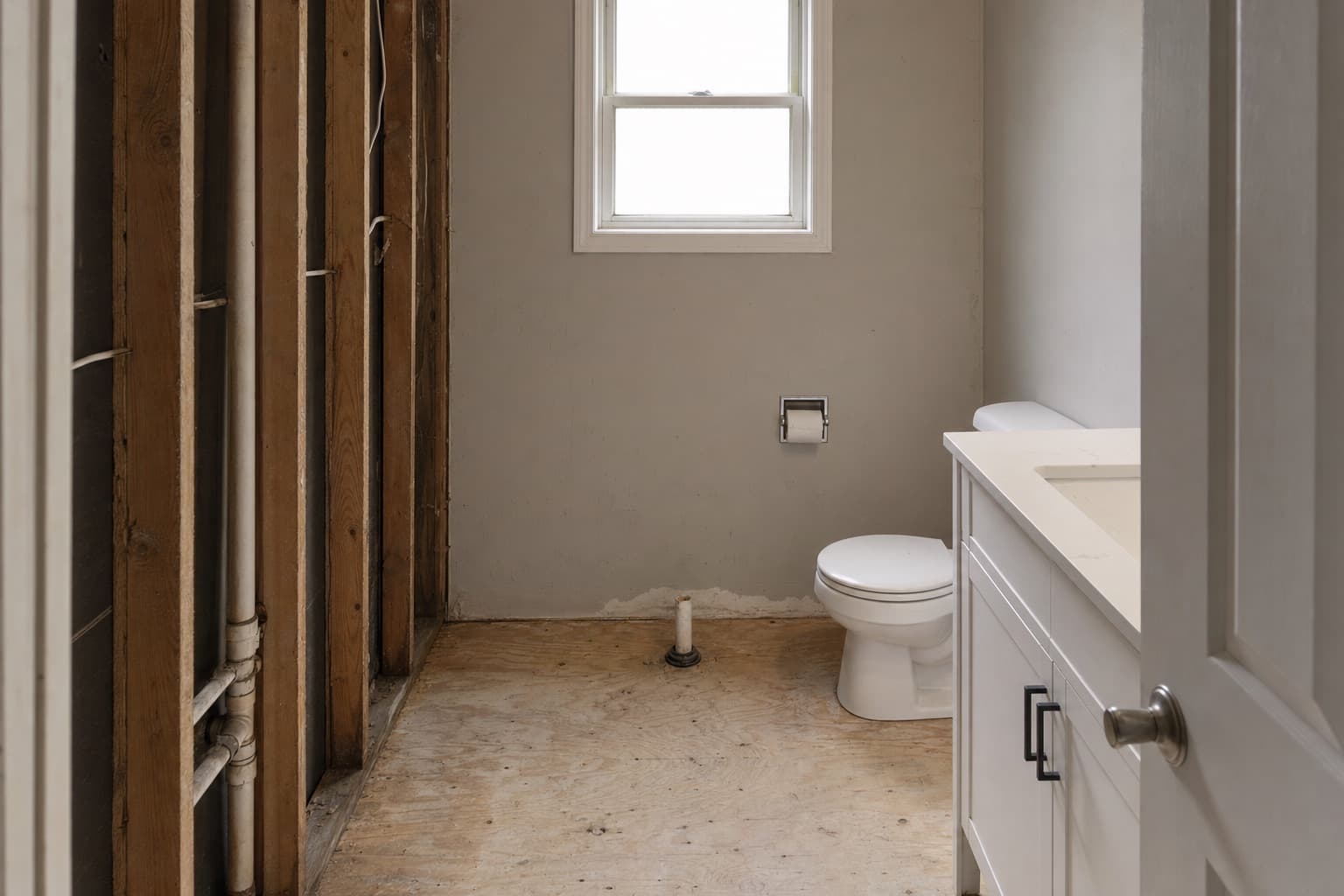 A small bathroom stripped for conversion, the floor bare to the subfloor with the drain pipe stubbed up and the wall studs exposed